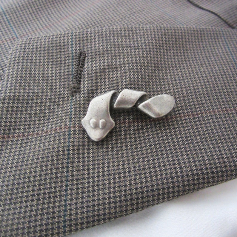 May include: A silver-toned metal lapel pin in a stylized, abstract design resembling a snake or ribbon. The pin is placed on a gray and brown patterned fabric, likely a suit jacket.