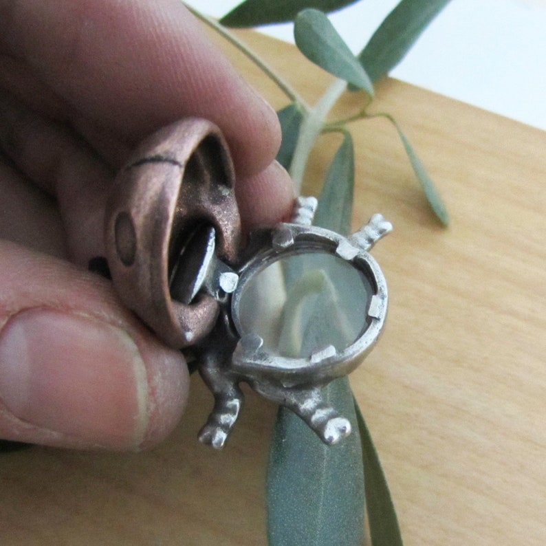 May include: A handcrafted, open-faced turtle pendant with a copper-colored shell and a silver-toned body. The turtle's shell opens to reveal a dark, oval-shaped stone. The pendant is held against a light wooden surface.