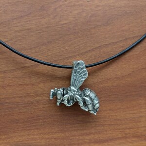 May include: A silver bee pendant necklace with a black leather cord. The bee is detailed with wings, legs, and antennae.