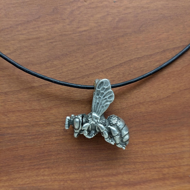 May include: A silver bee pendant necklace with a black leather cord. The bee is detailed with wings, legs, and antennae.