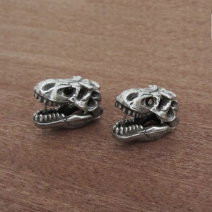 May include: Two silver-colored dinosaur skull earrings. The earrings are detailed with open jaws and visible teeth. The skulls are set against a warm, brown wooden surface.