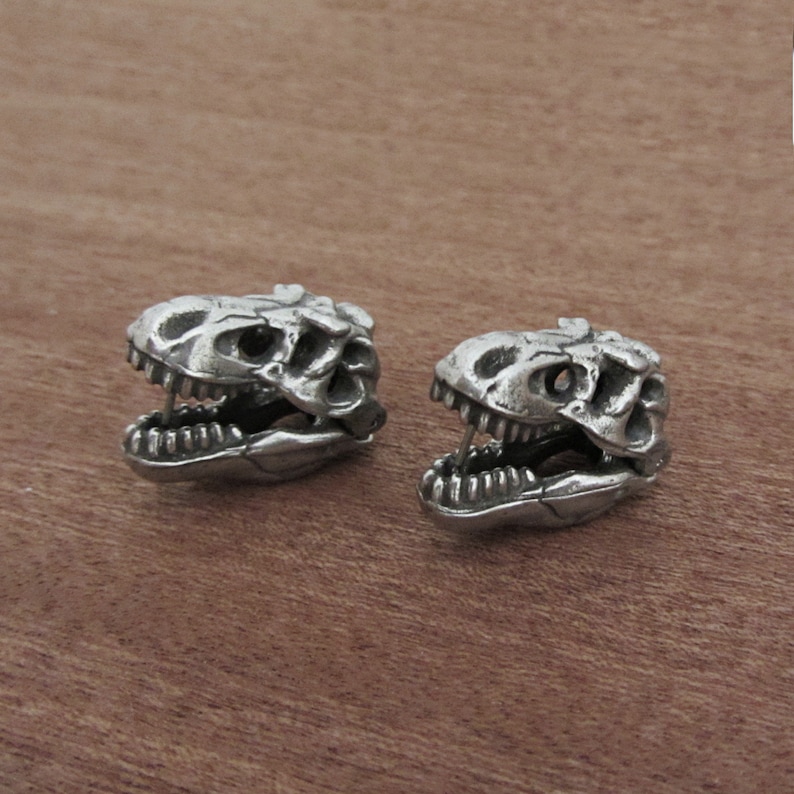 May include: Two silver-colored dinosaur skull earrings. The earrings are detailed with open jaws and visible teeth. The skulls are set against a warm, brown wooden surface.