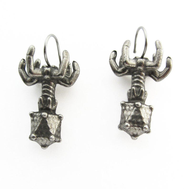 May include: A pair of silver earrings featuring a detailed design of a creature with multiple arms holding a geometric shape.