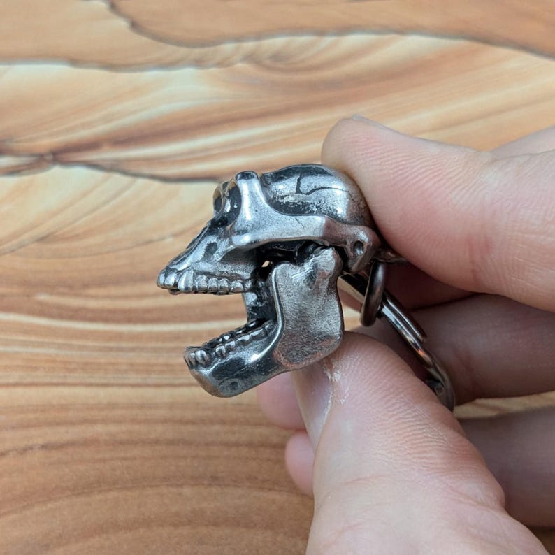 Puede incluir: Un llavero de metal plateado con un dise&ntilde;o detallado de cr&aacute;neo de primate con una mand&iacute;bula articulada en posici&oacute;n abierta. El cr&aacute;neo tiene dientes visibles, cuencas oculares y un acabado de superficie texturizado unido a una anilla met&aacute;lica.