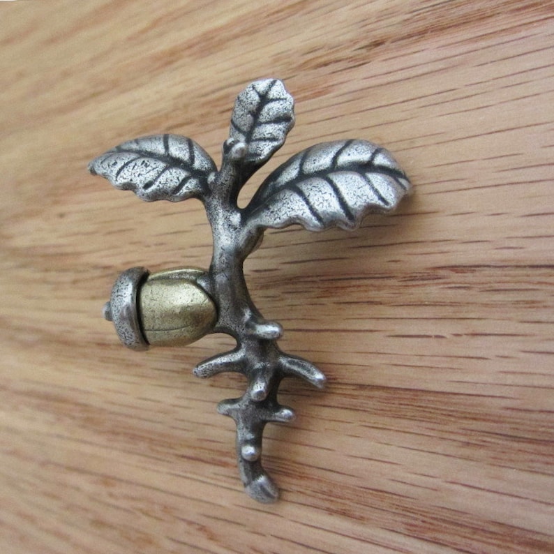 May include: A silver-toned metal acorn and oak leaf decorative element. The acorn has a gold-colored cap and is attached to a branch with three leaves. The piece is set against a wooden background.