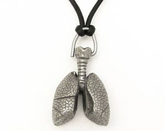 Puede incluir: Un collar con colgante plateado que presenta un diseño anatómico detallado de pulmones. El colgante está suspendido de un cordón negro. Los pulmones tienen textura y forma realista, con tráquea y bronquios visibles.