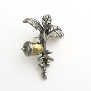 May include: A silver-toned metal brooch in the shape of a branch with leaves and an acorn. The acorn has a gold-colored body and a silver cap. The brooch has a pin on the back for attachment.