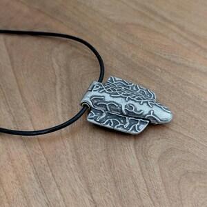 Puede incluir: Collar con colgante plateado y cord&oacute;n negro. El colgante presenta un dise&ntilde;o abstracto y texturizado con patrones detallados. El collar se muestra sobre una superficie de madera.