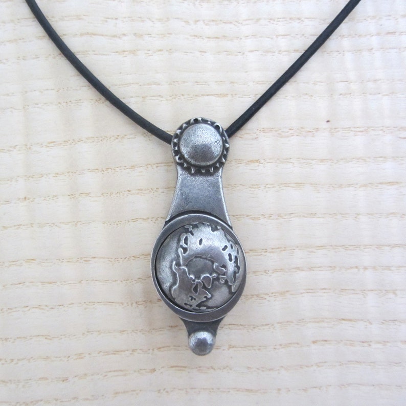 May include: A silver pendant necklace with a detailed globe design. The pendant is attached to a black cord.