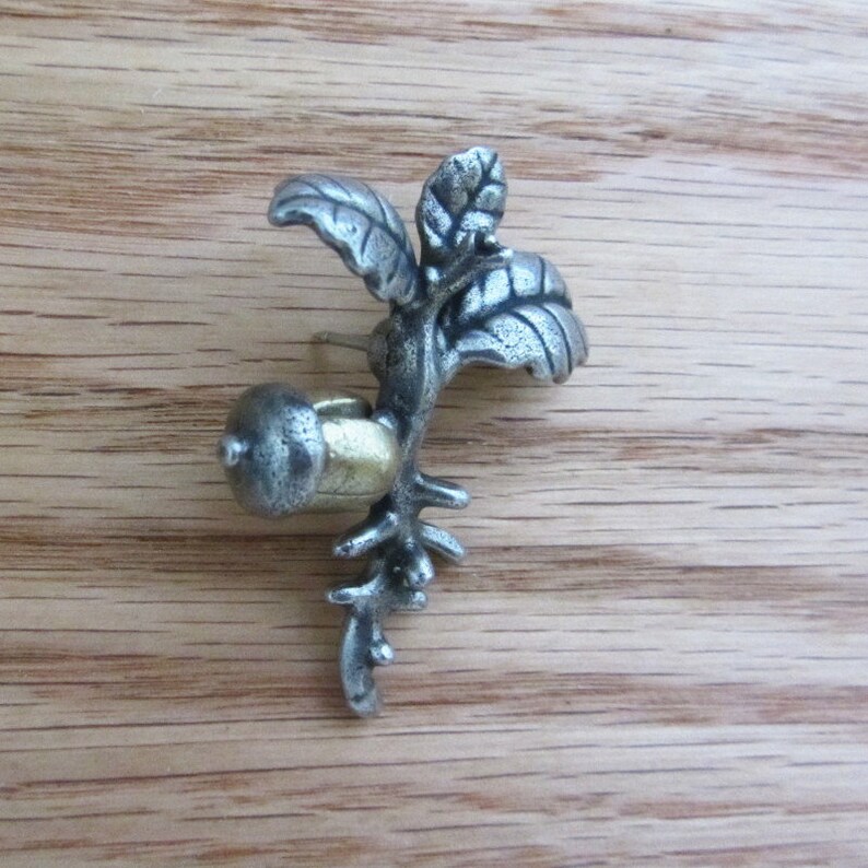 May include: A silver-toned metal earring in the shape of a branch with leaves and an acorn. The acorn has a gold-colored cap. The earring is set against a wooden background.