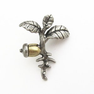 May include: A silver-toned metal brooch in the shape of a small tree with an acorn. The brooch features detailed leaves and a textured finish, with a gold-colored acorn. The design is nature-inspired.