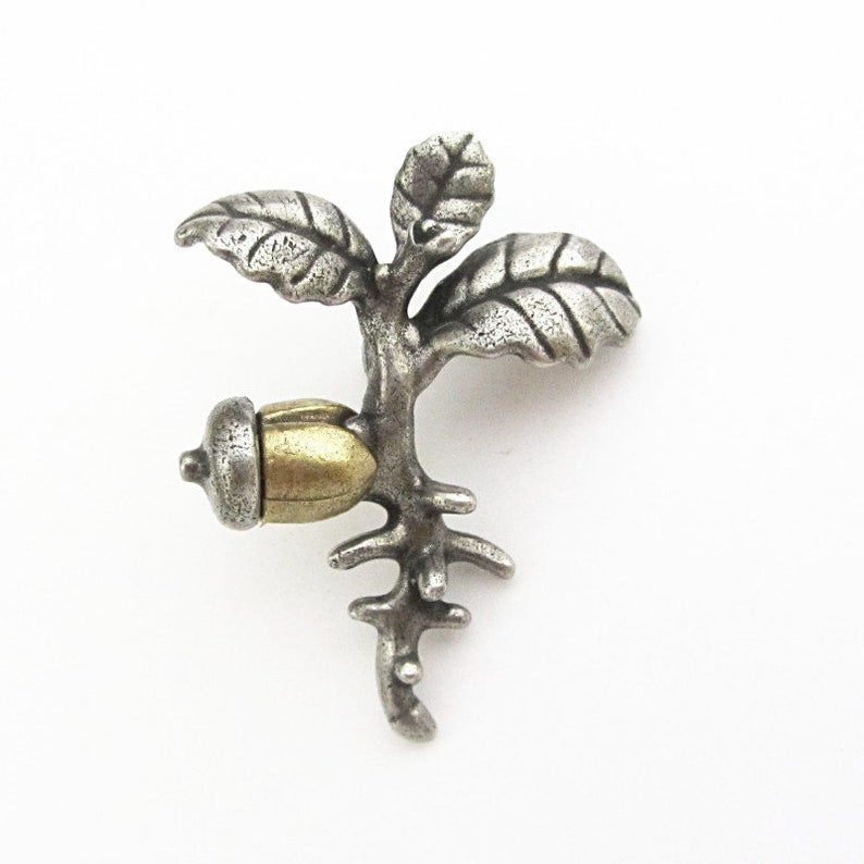 May include: A silver-toned metal brooch in the shape of a small tree with an acorn. The brooch features detailed leaves and a textured finish, with a gold-colored acorn. The design is nature-inspired.