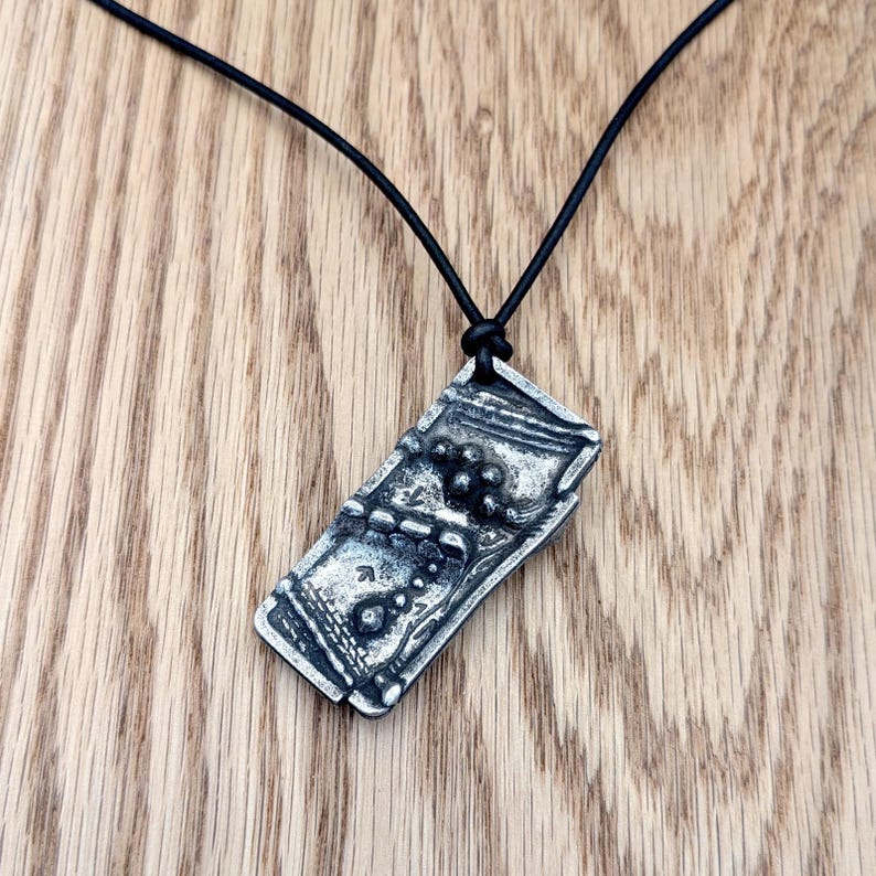 Op de afbeelding: Een rechthoekige zilveren hanger met een getextureerd oppervlak, hangend aan een zwarte koordketting. De hanger heeft een abstract ontwerp met verhoogde details en een donkere, geoxideerde afwerking. De ketting wordt gepresenteerd op een houten oppervlak.