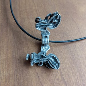 May include: A silver pendant necklace with a detailed, intricate design of a winged creature. The pendant is open and reveals a smaller, detailed figure inside. The necklace is on a black cord.