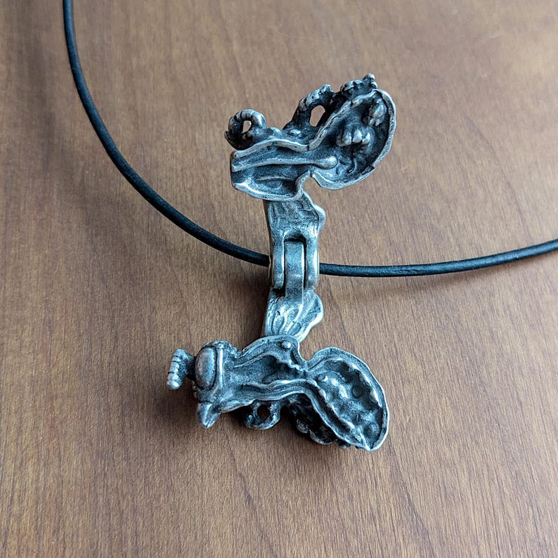 May include: A silver pendant necklace with a detailed, intricate design of a winged creature. The pendant is open and reveals a smaller, detailed figure inside. The necklace is on a black cord.