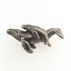 May include: A pewter-colored sculpture of two whales, one larger with a smaller whale riding on its back. The whales are detailed with textured surfaces and fins, set against a white background. The sculpture is a decorative item.