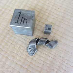 May include: A metal sculpture with a twisted design, next to a 1-inch cube and a 1 cm cube. The sculpture has a textured, aged appearance. The cubes are made of a similar metal.