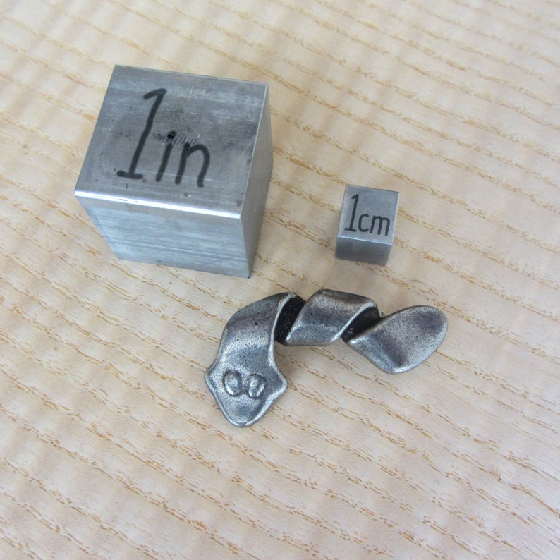 May include: A metal sculpture with a twisted design, next to a 1-inch cube and a 1 cm cube. The sculpture has a textured, aged appearance. The cubes are made of a similar metal.