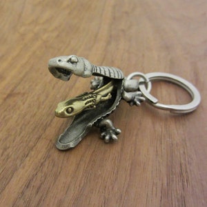 May include: A detailed metal keyring featuring a lizard design. The keyring has a silver-coloured outer lizard with a gold-coloured lizard inside. The keyring includes a silver-coloured ring.