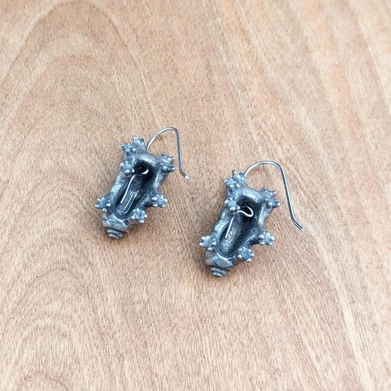 May include: A pair of silver-toned metal drop earrings featuring a textured, irregular shell-like shape with small protruding bumps and a hollow center. Each earring is attached to a thin, curved silver-toned wire hook for ear attachment.