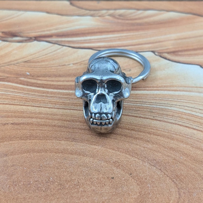 Puede incluir: Un llavero de metal plateado con un dije detallado en forma de cr&aacute;neo de primate. El cr&aacute;neo presenta arcos superciliares prominentes, cuencas oculares profundas, cavidad nasal y dientes visibles. Un anillo met&aacute;lico circular est&aacute; unido a la parte superior.