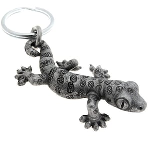 May include: A silver-coloured metal keyring featuring a detailed gecko design. The gecko has a textured body with dark spots and stripes, and is attached to a silver key ring. The reptile is approximately 10 cm long.