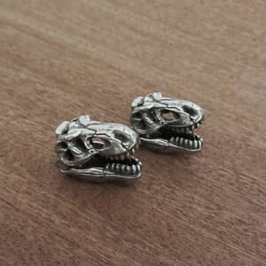 May include: Two silver-colored dinosaur skull beads. The beads are detailed with visible teeth and skull features. The skulls are positioned on a wooden surface, showcasing the intricate design of the prehistoric theme.