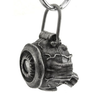 May include: A detailed, antique-style silver-colored pendant with a unique, mechanical design. The pendant features a circular element with a central eye-like detail, and is suspended from a silver-colored ring. The overall aesthetic suggests a steampunk or industrial theme.