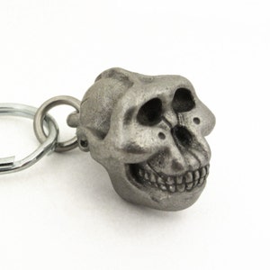 May include: A detailed, metallic keychain charm shaped like a primate skull. The skull has a detailed texture, visible teeth, and eye sockets. The keychain ring is silver and attached to the skull's head.