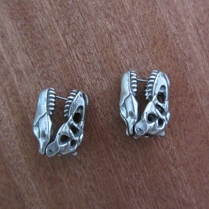 May include: A pair of silver-toned earrings. Each earring is shaped like a dinosaur jaw, with detailed teeth and bone structure. The earrings are designed to be worn as a pair, with a post for piercing. The earrings are set against a brown background.
