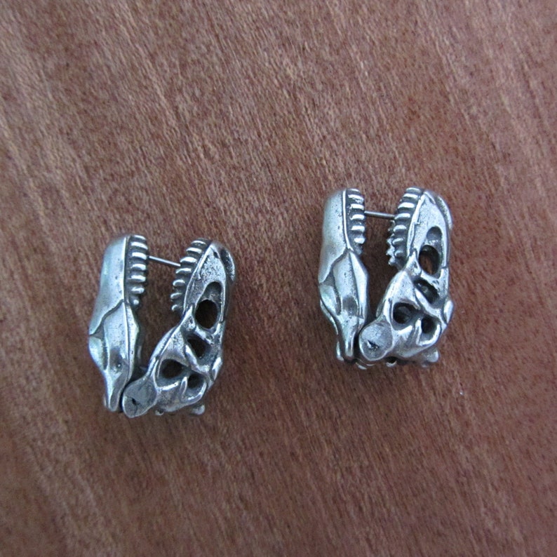 May include: A pair of silver-toned earrings. Each earring is shaped like a dinosaur jaw, with detailed teeth and bone structure. The earrings are designed to be worn as a pair, with a post for piercing. The earrings are set against a brown background.