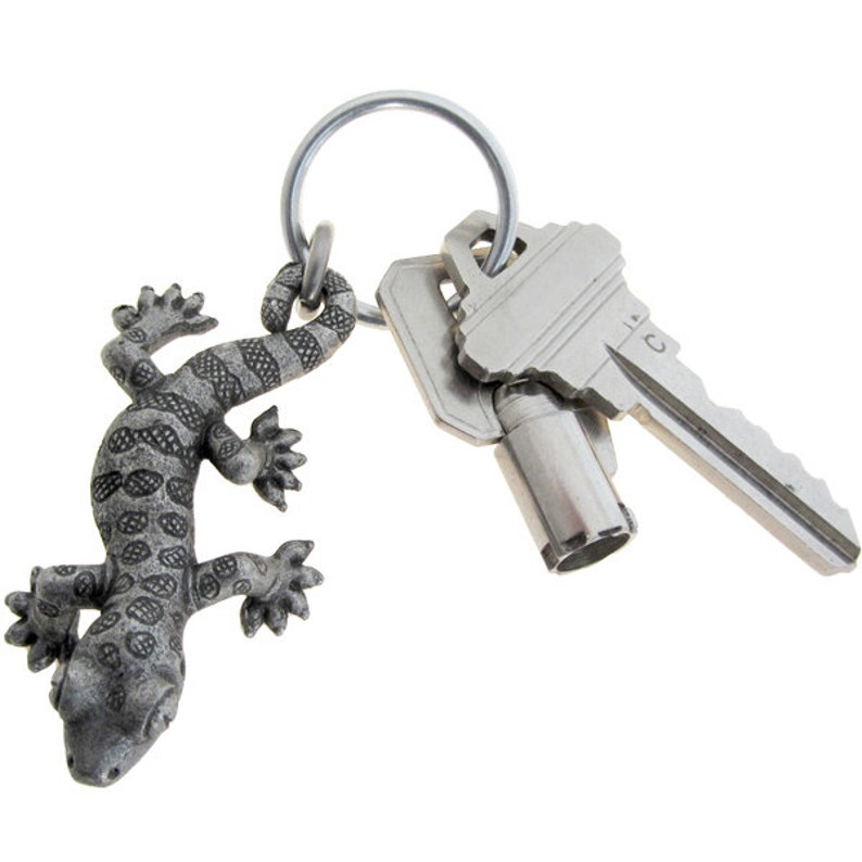 May include: A silver key ring featuring a detailed gecko charm and two keys. The gecko charm has a textured, spotted pattern. The keys are metal and attached to the ring. The key ring is set against a white background.