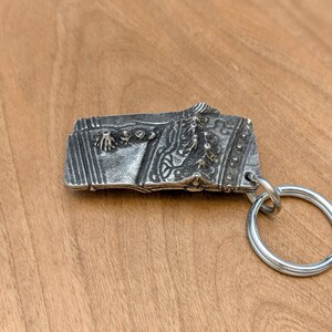 May include: A silver-colored rectangular keychain with intricate embossed designs. The keychain features a detailed relief of a historical scene, attached to a large, round silver key ring. The background is a light brown wooden surface.