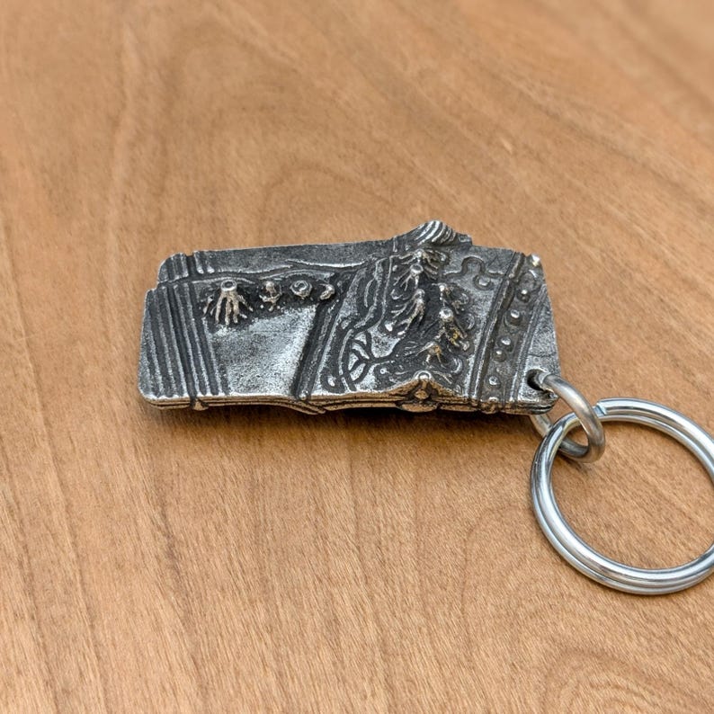 May include: A silver-colored rectangular keychain with intricate embossed designs. The keychain features a detailed relief of a historical scene, attached to a large, round silver key ring. The background is a light brown wooden surface.