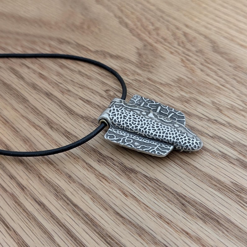 Puede incluir: Un collar con colgante plateado y cord&oacute;n negro. El colgante tiene un dise&ntilde;o texturizado y en capas con una mezcla de patrones. El collar se muestra sobre una superficie de madera.