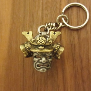 May include: A detailed keychain featuring a samurai mask pendant. The mask is a combination of silver and gold tones, with intricate details. The keychain includes a metal ring and chain for attaching to keys or bags.