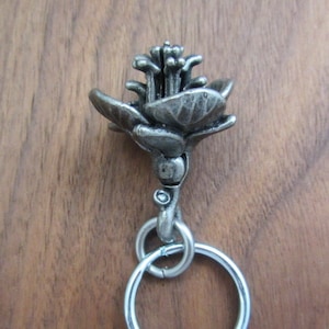 May include: A silver-toned metal keychain featuring a detailed flower design. The flower has multiple petals and a textured surface. The keychain includes a split ring for attaching keys. The background is a wooden surface.