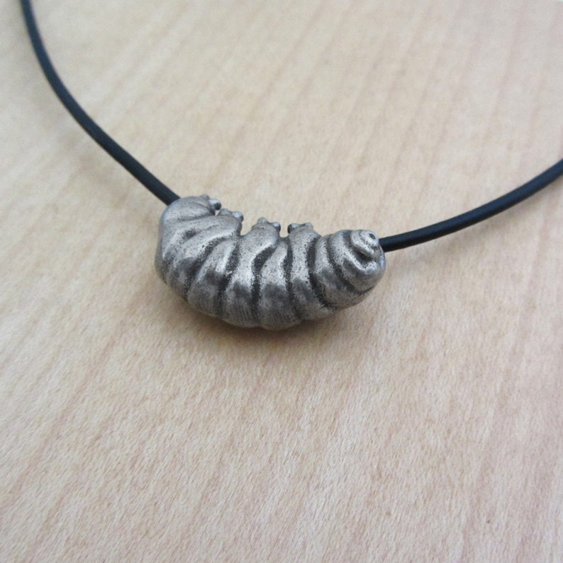 May include: A silver-toned caterpillar pendant necklace. The pendant is detailed with segmented body ridges and is strung on a black cord. The necklace is a unique piece of jewelry.