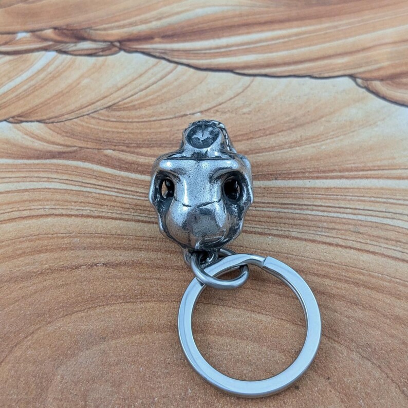 Puede incluir: Un llavero de metal plateado con un dije de calavera en tres dimensiones. La calavera tiene cuencas oculares profundas y una cavidad nasal detallada, unida a una argolla circular para llaves. La superficie met&aacute;lica tiene un acabado oscuro.
