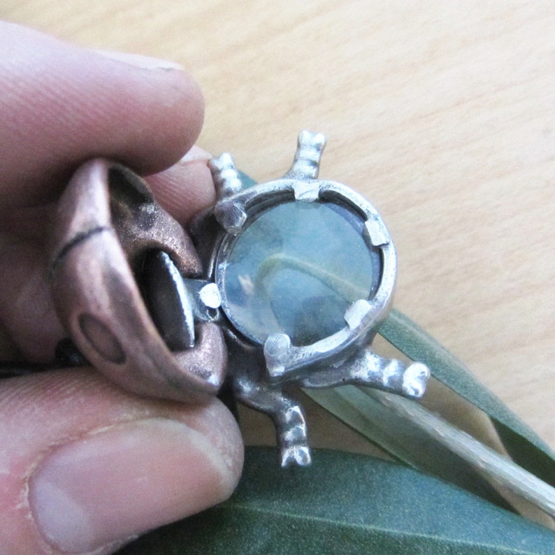 May include: A handcrafted silver ring with a large, round, pale blue gemstone. The ring has a unique, organic design with decorative elements. The ring is held by a person's fingers, showcasing the intricate details and craftsmanship.