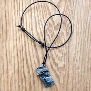 Op de afbeelding: Een zilverkleurige hangerketting met een zwart koord. De rechthoekige hanger heeft een abstract ontwerp met verhoogde details en een getextureerd oppervlak. Het koord is verstelbaar met een schuifknoop. De ketting ligt op een houten oppervlak.