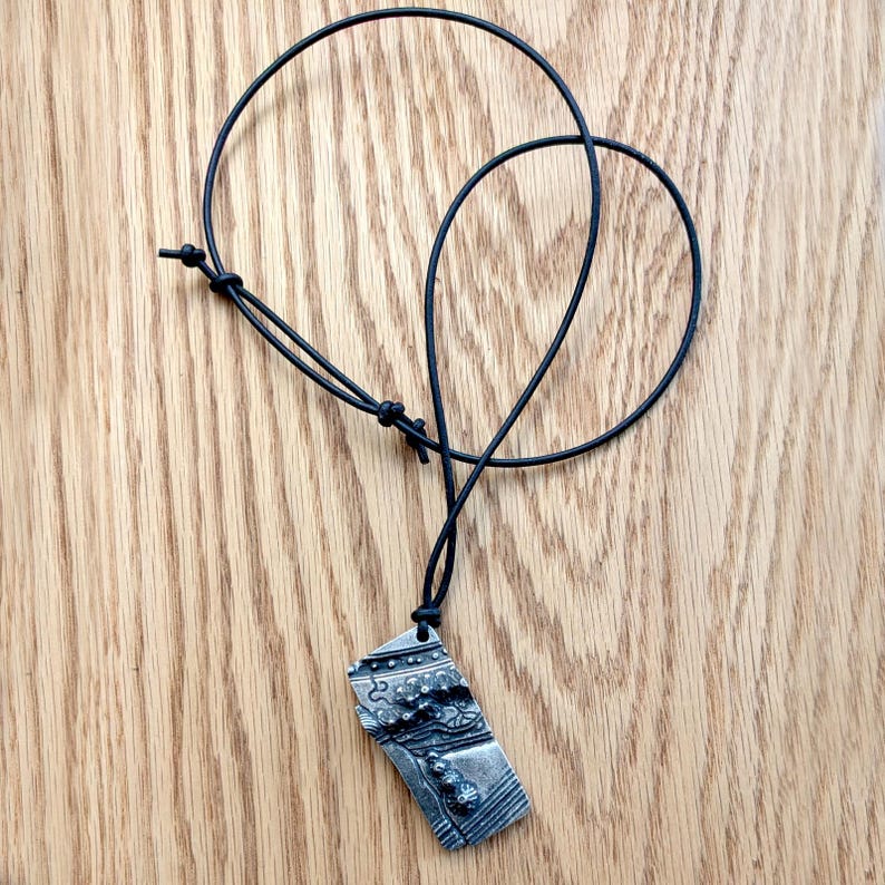 Op de afbeelding: Een zilverkleurige hangerketting met een zwart koord. De rechthoekige hanger heeft een abstract ontwerp met verhoogde details en een getextureerd oppervlak. Het koord is verstelbaar met een schuifknoop. De ketting ligt op een houten oppervlak.