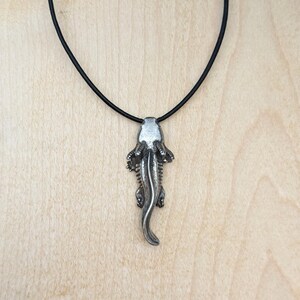 May include: A silver-colored pendant in the shape of an alligator or crocodile, suspended from a black cord necklace. The pendant has detailed features, including the head, legs, and tail. The necklace is displayed against a light-colored wooden background.
