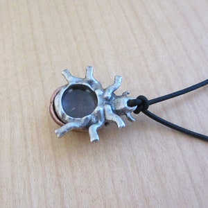 May include: A silver-toned pendant with a dark, circular stone center and a sunburst design. The pendant is attached to a black cord. The pendant has a rustic, handcrafted appearance.