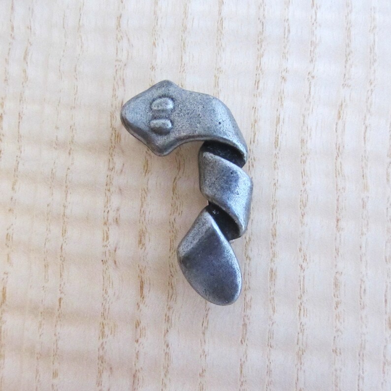 May include: A pewter-colored, stylized snake-shaped cabinet pull. The pull features a snake head with two eye indentations and a spiraling body. The hardware is designed to be mounted on a wooden surface.