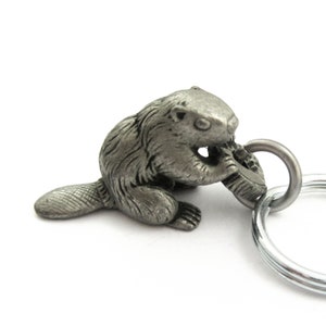 May include: A silver-colored metal keychain featuring a detailed beaver figurine. The beaver is depicted gnawing on a piece of wood, with a textured fur design. The keychain includes a split ring for attaching keys.