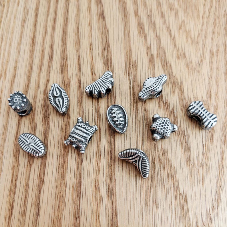 May include: A collection of ten small, silver-toned metal beads with various embossed textures and geometric patterns. Each bead features a unique shape, including oval, rectangular, and curved forms, all displaying a weathered, oxidized finish.