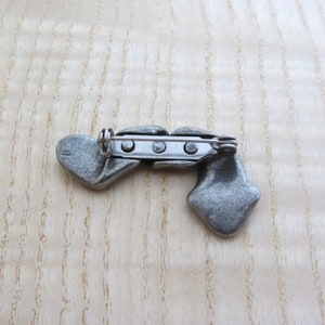 May include: A silver-toned metal brooch with a unique design. The brooch features two heart-shaped elements and a diamond-shaped element, connected by a central bar with a pin closure. The brooch is set against a light wood grain background.