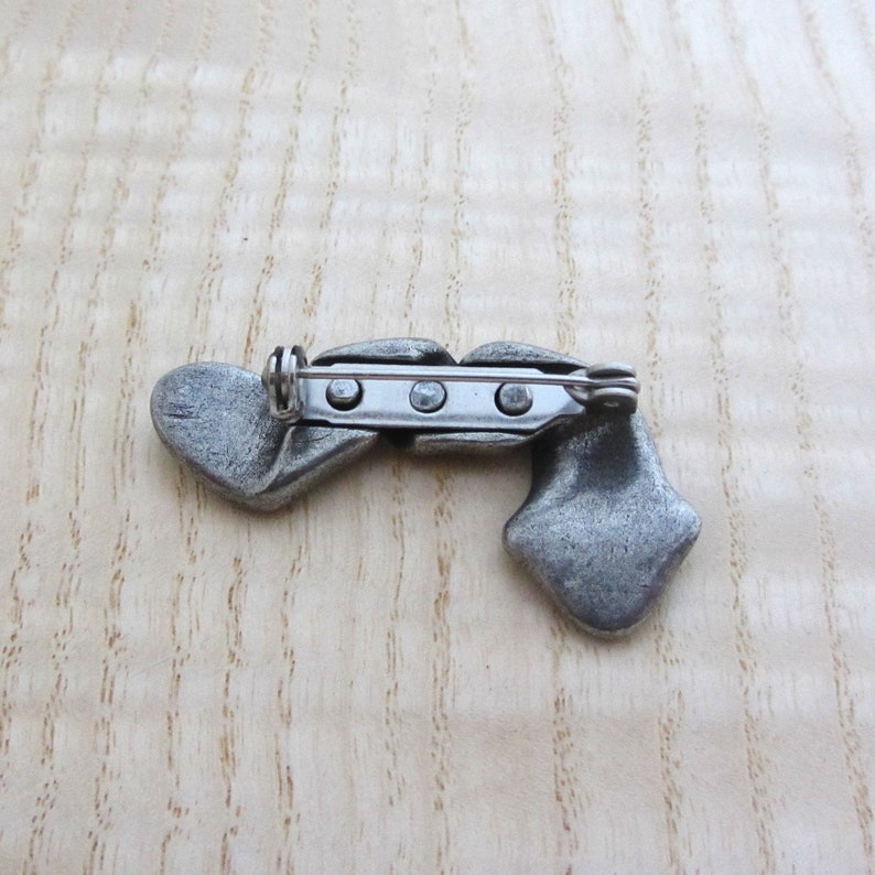 May include: A silver-toned metal brooch with a unique design. The brooch features two heart-shaped elements and a diamond-shaped element, connected by a central bar with a pin closure. The brooch is set against a light wood grain background.