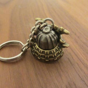 May include: A detailed keychain featuring a miniature helmet design. The helmet is made of metal with a silver and gold finish, attached to a chain and keyring. The helmet has intricate detailing and a vintage aesthetic.